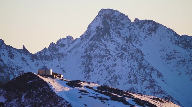 A small mountain hut stands on a snowy ridge, bathed in the warm, golden light of the setting sun. The rugged, snow covered peaks of the surrounding mountains dominate the background, creating a majes