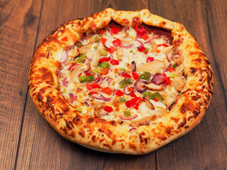 A cooked pizza with a lot of toppings including chicken, peppers, and onions. The pizza is sitting on a wooden table. Fine Italian style meal popular world wide. Rich dough base.