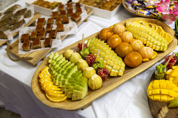  carved fresh tropical fruit, including precisely cut yellow mango, orange slices, clementines, and kiwi, arranged beautifully on a wooden platter as part of a larger dessert buffet.