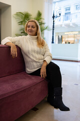 Fototapeta premium A blonde woman in a knitted sweater is sitting on a sofa in a modern interior. She is wearing a white sweater and black trousers.
