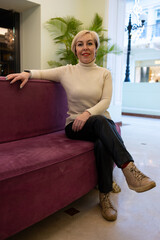 A blonde woman in a knitted sweater is sitting on a sofa in a modern interior. She is wearing a white sweater and black trousers.