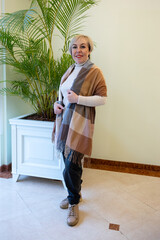 A blonde woman in a knitted sweater is sitting on a sofa in a modern interior. She is wearing a white sweater and black trousers.