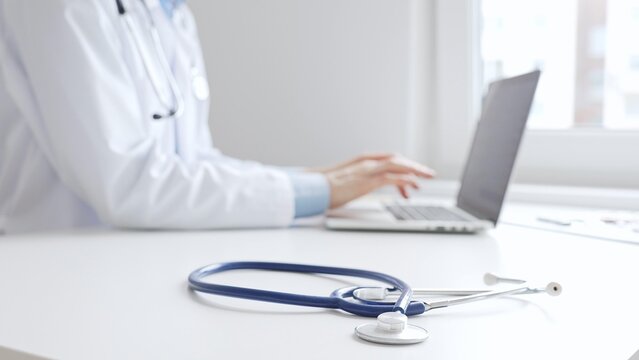 Closeup of blue stethoscope is lying on the table near female doctor working on laptop, highlighting digital tools transforming healthcare diagnostics and patient documentation. Medicine concept - Powered by Adobe