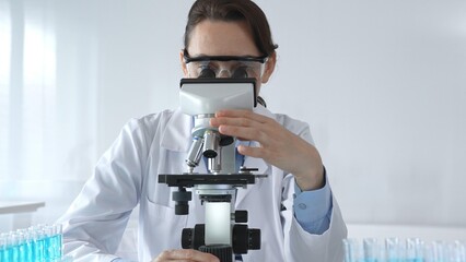 Female scientist wearing lab coat and protective glasses using microscope, analyzing samples in test tubes in modern laboratory, conducting scientific research