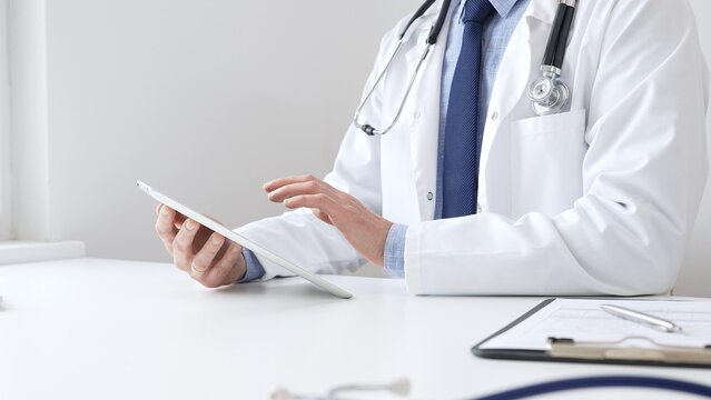 Doctor man wearing white medical coat and stethoscope using digital tablet while sitting at desk in medical office, with medical chart and pen in the foreground. Medicine concept - Powered by Adobe