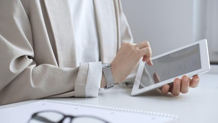 Professional woman working on tablet at desk, checking digital content with notebook and glasses nearby, focused on online tasks. Business people concept
