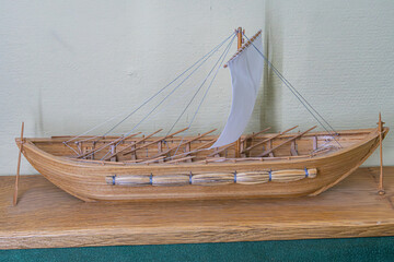 Ancient galley model wood, multiple masts, square sails, oars, flat bottom hull, intricate details rigging, oarlocks, textured surface, wooden stand, angled perspective