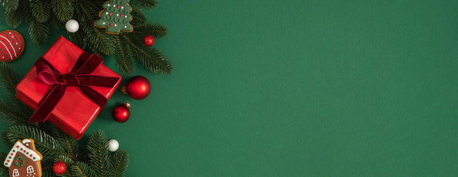 Christmas background: festive green backdrop with fir tree branches, gift box with velvet ribbon, gingerbread cookies and red baubles. Holiday border, copy space, flat lay, top view, New Year, winter.