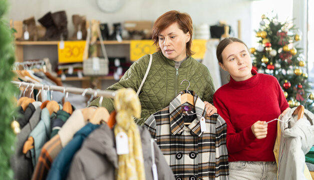 Adult woman and teenage girl choosing warm plaid coat or down jacket in clothing store decorated for christmas