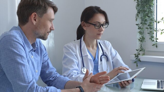 Female doctor using a digital tablet for explaining test results to a male patient during a consultation in a bright, modern medical office. Medicine concept