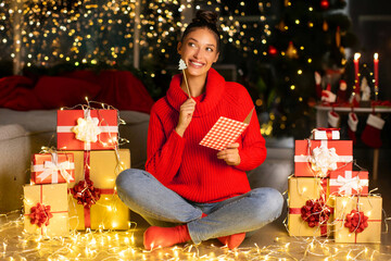 Dreamy young lady in red sweater thinking and writing list with gifts, sitting among wrapped gift...