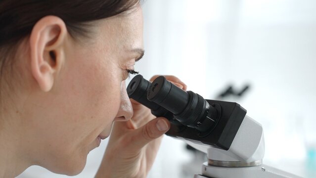 Female scientist peering into microscope, analyzing biological sample with precision in sterile laboratory research environment. Medicine and science concept