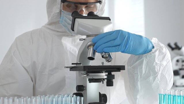 Medical virologist in protective hazmat gear examining viral samples under a microscope while conducting research in a sterile laboratory environment - Powered by Adobe