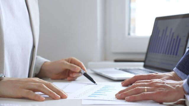 Financial analysts reviewing laptop screen and printed documents, pointing at data charts and discussing investment strategies together in modern office setting. Business people concept