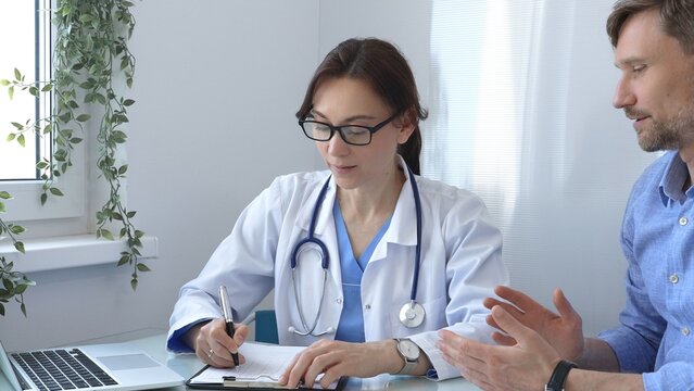 Female doctor consulting with patient in medical office using laptop and filling out paperwork to provide healthcare advice and support, discussing treatment options in a professional setting. Medical - Powered by Adobe