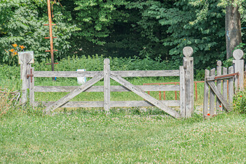 Ancient Central European village, lush green field, weathered wooden gate, planks and rails, brown tones, vibrant contrast, well-tended grass, diverse trees, overcast day or diffuse cloud cover, peac