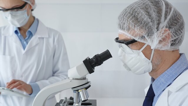 Senior male scientist wearing protective mask, glasses and hairnet using microscope with young female colleague assisting, holding tablet and taking notes in laboratory. Medicine and science concept
