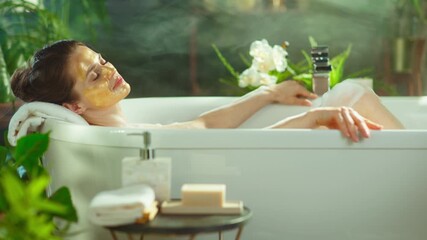 Woman with golden facial mask relaxes blissfully in a bubble bath adorned with flowers, eyes closed. Bright, plant-filled bathroom with soft lighting & nearby skincare captures luxurious self-care. - Powered by Adobe