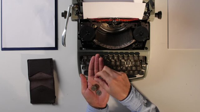 Hands carefully counting a few coins, signifying financial struggle and the pursuit of income through creative work, placed above a vintage typewriter and blank paper on a minimalist desk