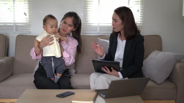 A woman is holding a baby while sitting on a couch. Another woman is sitting next to them, looking at a tablet. The scene appears to be a casual and friendly gathering between the two women