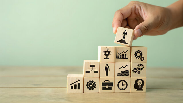 Hand placing wooden block with winner podium icon atop ascending staircase of success symbols including hierarchy person graph gear growth briefcase clock brain on wooden table against soft green back