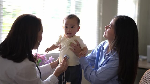 A woman is holding a baby and a doctor is checking the baby's heartbeat. The baby is wearing a white shirt