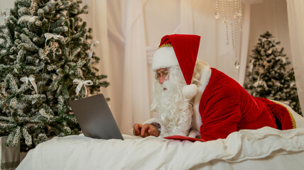 Santa Claus using a laptop computer on Christmas morning in a festive bedroom setting