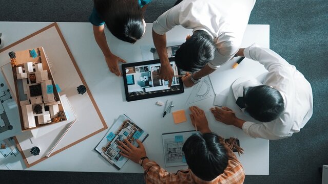 Top down view of architect engineer team discussing and point at tablet display construction at table with blueprint and house model at meeting. Diverse designer planning design house. Alimentation. - Powered by Adobe