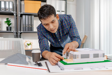 A young architect works with building plans and a house model in the office, reviewing blueprints, marking details, and analyzing design structures with focus and creativity.