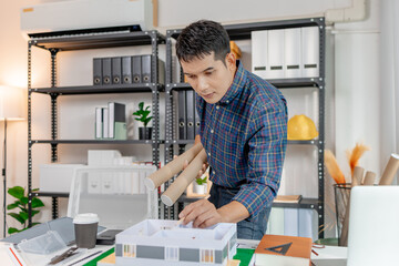 A young architect is working at his desk, reviewing blueprints, using a calculator, and checking a house model. He focuses on precision and detail while planning construction projects.