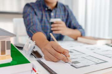 A young architect is working at his desk, reviewing blueprints, using a calculator, and checking a house model. He focuses on precision and detail while planning construction projects.