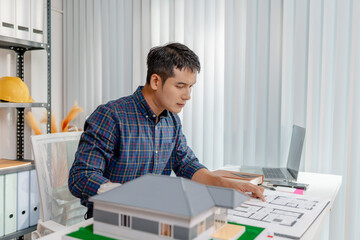 A young architect is working at his desk, reviewing blueprints, using a calculator, and checking a house model. He focuses on precision and detail while planning construction projects.