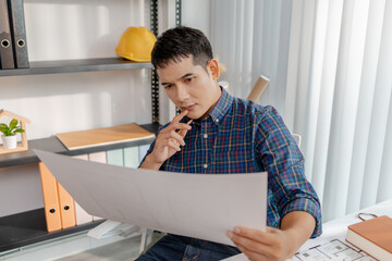 A young architect is working at his desk, reviewing blueprints, using a calculator, and checking a house model. He focuses on precision and detail while planning construction projects.