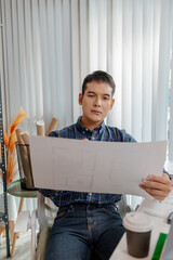 A young architect is working at his desk, reviewing blueprints, using a calculator, and checking a house model. He focuses on precision and detail while planning construction projects.