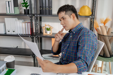 A young architect is working at his desk, reviewing blueprints, using a calculator, and checking a house model. He focuses on precision and detail while planning construction projects.