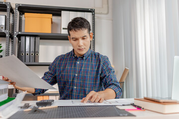 A young architect is working at his desk, reviewing blueprints, using a calculator, and checking a house model. He focuses on precision and detail while planning construction projects.