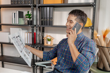 A young architect is standing at his desk while discussing a project on the phone, carefully reviewing construction plans with focus and attention in a modern office environment.