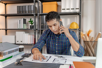 A young architect is standing at his desk while discussing a project on the phone, carefully reviewing construction plans with focus and attention in a modern office environment.