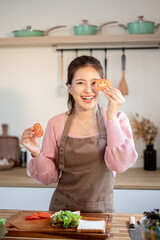 Happy asian woman wearing apron holding tomato slices making a sandwich at kitchen cooking counter.
