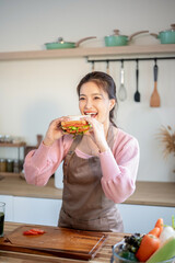 Happy asian woman wearing apron holding and eating sandwich after making at kitchen cooking counter.
