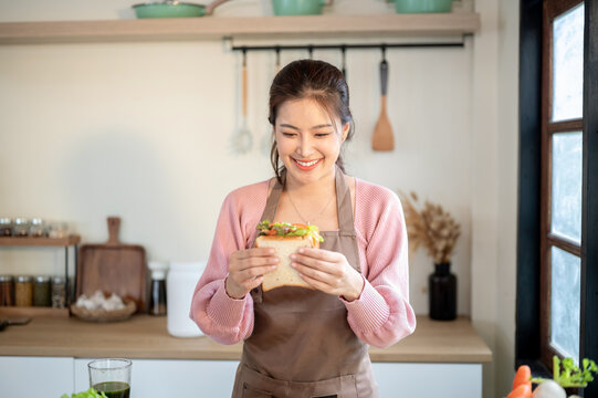 Happy asian woman wearing apron holding and looking at sandwich she makes at kitchen cooking counter