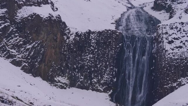 A view of the Terskol waterfall cascading down a snowy, rocky mountain face in the North Caucasus region of Russia. The powerful flow of water is framed by a pristine winter landscape, with snow and i