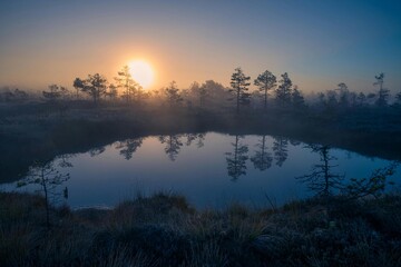 Beautiful sunrise with orange and blue sky over a calm river and forest, showing a reflection in the water and mist around the trees