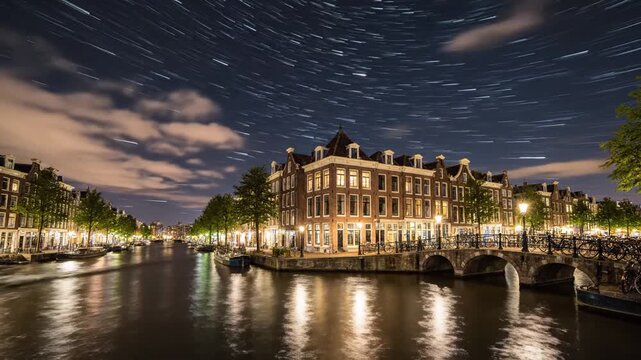 Night Canal View In Amsterdam Netherlands Featuring Illuminated Buildings Reflecting On Water Under A Sky With Streaking Stars And Wispy Clouds For Adobe Stock
