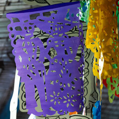 A purple paper lantern with a skeleton on it hangs on a wall. The lantern is decorated with flowers. Mexican Day of the Dead tradition, offerings, food, colors, decorations, and papel picado, La Catri