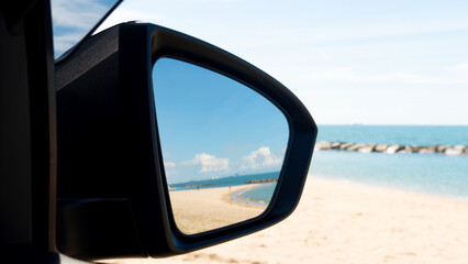 Mirror wing side of car in side with sand beach under blue sky. Outside of car with clear blue sky and turquoise sea with coastal rock barrier.