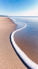 A gentle wave with white foam rolls onto a sandy beach, meeting the calm ocean under a bright blue sky.