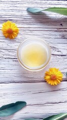 A clear jar of yellow balm is placed on a rustic white wooden surface, adorned with vibrant calendula flowers and green sage leaves.