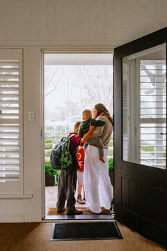 Mother holding toddler son in her hip leading older son with backpack out of the doorway