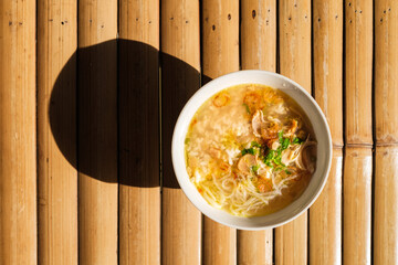 Overhead shot of nusantara indonesian food calles soto filled with chicken noodle soup in a white bowl, placed on a bamboo mat with a shadow.